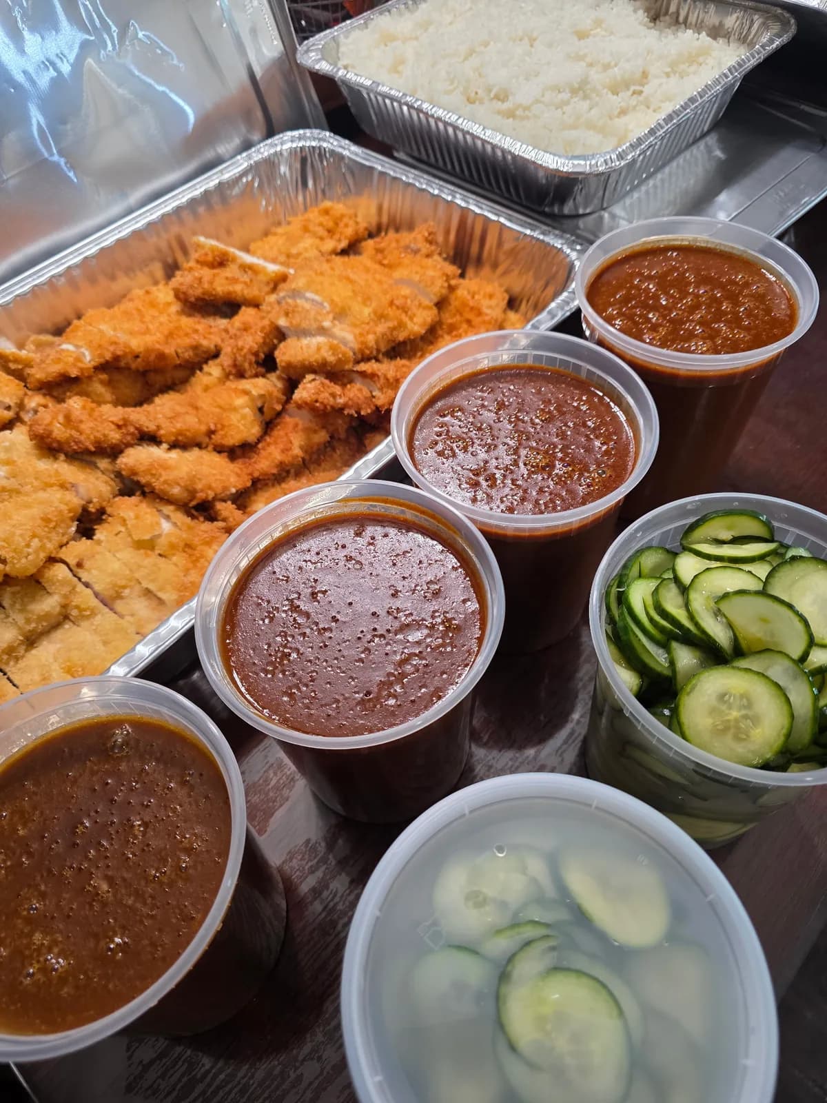 Catering spread with breaded chicken cutlets, steamed rice, sauces, and sliced cucumbers